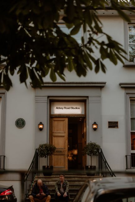 The image shows the exterior of a building with a white facade and a wooden doorway, labeled with a sign above the entrance that reads 'Abbey Road Studios.' On either side of the entrance, there are t
